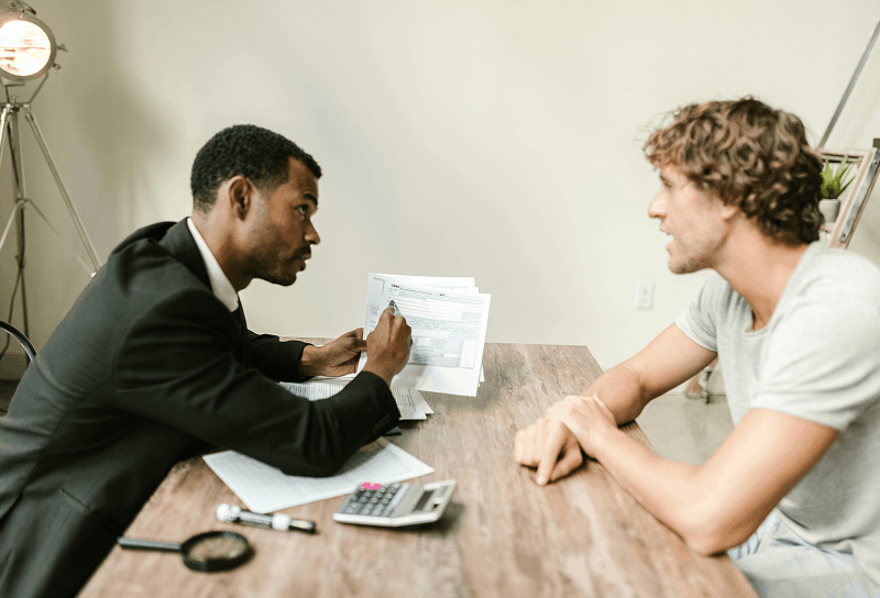 Consultant reviewing documents and discussing business or financial matters with a client across a desk in a professional office setting.