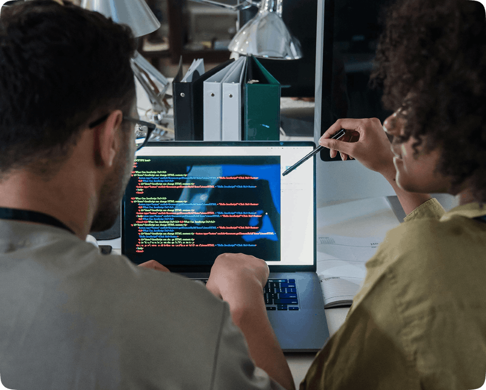Two developers collaborating on a laptop displaying programming code in a modern office workspace, representing teamwork in software and web development.
