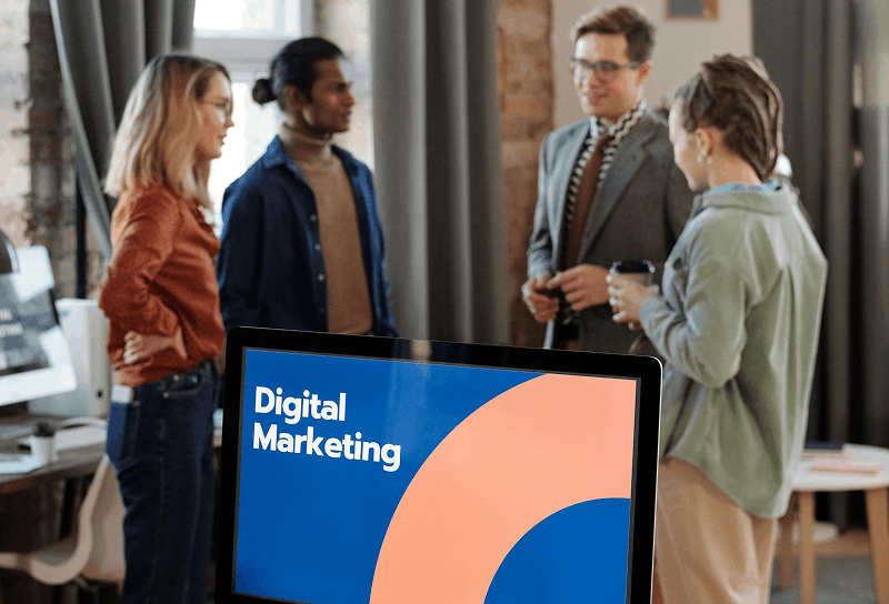 Marketing professionals discussing strategy in a modern office workspace with a computer screen displaying digital marketing branding in the foreground.